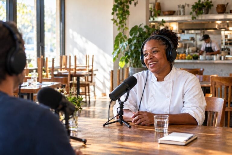 Restaurant owner in a chef's jacket recording a podcast at a table in her dining room